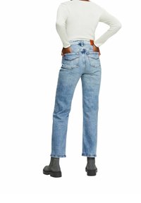 Jeans de mezclilla azul claro con corte recto, que cuentan con dos bolsillos traseros y un detalle de cuero marrón en la cinturilla. Combinados con botas negras hasta el tobillo.