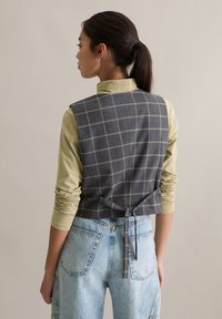 Szara kamizelka w kratę z detalem wiązania z tyłu, noszona na jasnzie zielonej koszuli z długim rękawem. Stylizację uzupełniają jasnoniebieskie jeansy z wysokim stanem.