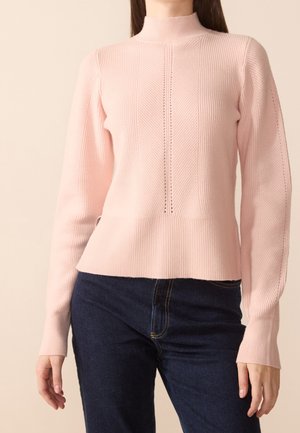 Vrouw die een lichtroze geribbelde gebreide trui draagt met lange mouwen en een hoge hals, gecombineerd met donkerblauwe spijkerbroek.