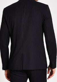 Mörk marinblå kavaj med smala vita strepor, strukturerade axlar, enkel vent och knäppta manschetter, tillverkad av ett slätt tyg.