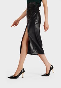 Gonna midi nera in pelle con alto spacco, abbinata a décolleté nere a punta con accentuazioni dorate e design a stiletto.