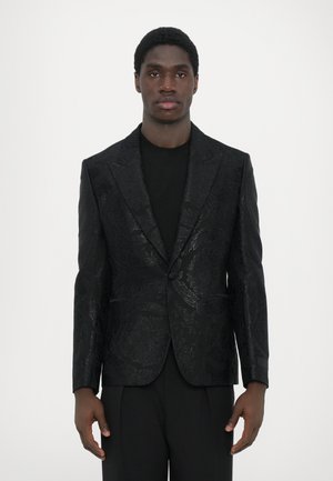 Schwarz gemusterter Blazer mit glänzender Blumenstruktur, einreihiger Knopfverschluss und Spitzrevers, getragen über einem schwarzen Oberteil. Maßgeschneiderte Passform.