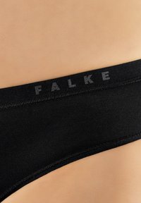 Zbliżenie na czarny pas z logo „FALKE” noszony na jasnej skórze.