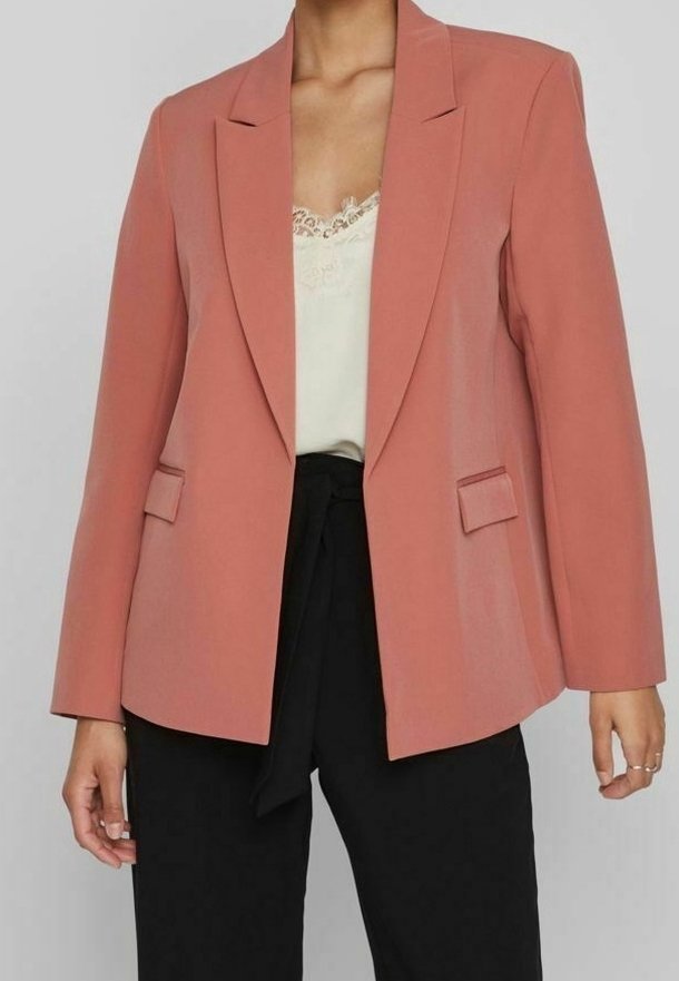 Femme portant un blazer rose saumon sur un caraco blanc bordé de dentelle et un pantalon noir taille haute avec ceinture à nouer, debout devant un fond uni.