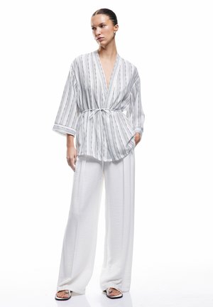 Haut blanc de style kimono avec des rayures verticales noires et une taille à nouer, associé à un pantalon blanc large. Texture lisse et coupe décontractée.