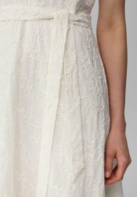 Robe en tissu blanc ornée de broderies complexes avec des motifs floraux tourbillonnants. Comprend une ceinture à nouer à la taille, avec une finition texturée.