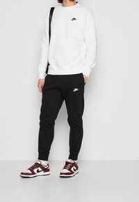 Bílá mikina s černým logem Nike, spárovaná s černými kalhotami s bílým logem. Dokončeno vínovými a bílými teniskami.