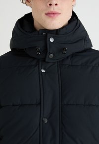 Chaqueta acolchada negra con cuello alto, que presenta un cierre frontal con cremallera, botones a presión negros y un cordón ajustable en el cuello.