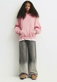 Ung person med lockigt hår som bär en rosa huvtröja, vida urtvättade gråjeans och tanfärgade mockasiner som står mot en vit vägg.
