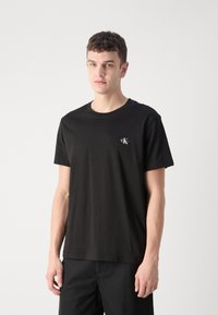 Camiseta de algodón negra con un cuello redondo, mangas cortas y un pequeño logo blanco en el lado izquierdo del pecho. Textura suave, corte relajado.
