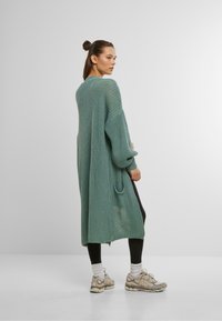 Long cardigan tricoté en vert clair avec un tissu texturé, des manches amples et une poche. Associé à des leggings noirs et des baskets beiges.