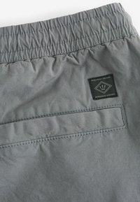 Pantaloni scurți gri cu talie elastică, având un eticheta aplicată și un buzunar pe spate. Materialul are o textură netedă cu detalii de cusătură vizibile.