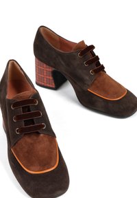 Brązowe zamszowe buty o dwukolorowym wzorze, z ciemniejszą brązową cholewką, pomarańczowymi akcentami i kratkowanym brązowym obcasem.