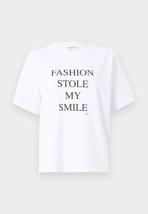 Vrouw met kort donker haar, gekleed in een wit grafisch T-shirt, donkere high-waisted wijde jeans, zwarte riem en zwarte open-toe hakken, staand tegen een witte achtergrond.