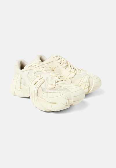 Cream-colored athletic shoes featuring a textured mesh upper, chunky rubber sole, and distinct sculpted accents on the toe and sides.