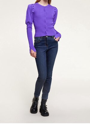 Woman wearing a fitted purple long-sleeve top with puffed shoulders, dark blue jeans, and black lace-up ankle boots standing with one hand in pocket.