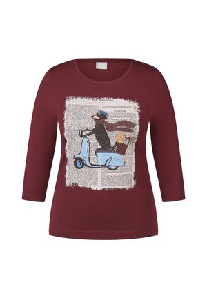 Frau trägt eine Jeansjacke über einem bedruckten bordeauxfarbenen Shirt, bordeauxfarbene Lederhose und bordeauxfarbene Sneakers mit weißen Streifen, steht vor einem schlichten Hintergrund.