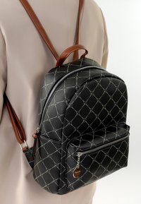 Musta kuvioitu reppu, jossa on ruskeat hihnat, puettu vaaleanruskeiden vaatteiden päälle, etutaskussa vetoketju ja hopeanväriset vetoketjun vetimet.