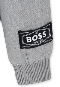 Tessuto grigio testurizzato con polsini a costine, con un'etichetta nera che recita "BOSS" con "seventy" in bianco e ulteriori dettagli di branding.