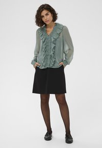 Groene, doorzichtige blouse met een gerimpelde voorkant en lange mouwen, gecombineerd met een zwarte rok en zwarte instapschoenen, wat een gelaagde look creëert.