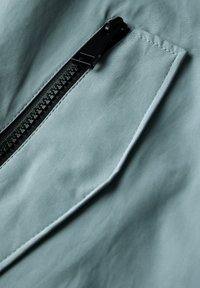 Tissu bleu clair avec une poche zippée dotée d'une fermeture éclair noire et d'une tirette texturée. Texture lisse avec des bords surpiqués et une finition soignée.