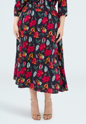 Kobieta w czarnej midi spódnicy i topie z jaskrawym czerwonym i żółtym kwiatowym wzorem, nosząca złote, odkryte na palce szpilki, stoi z rękami wzdłuż ciała.