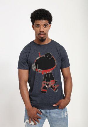 Ung mand med kort krøllet hår bærer en mørk t-shirt med en tegneseriebjørn i røde og sorte seler samt lyse revnede jeans.
