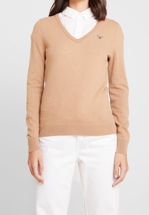 Personne portant un pull beige en V par-dessus une chemise blanche boutonnée avec un pantalon blanc, debout devant un fond gris clair uni.