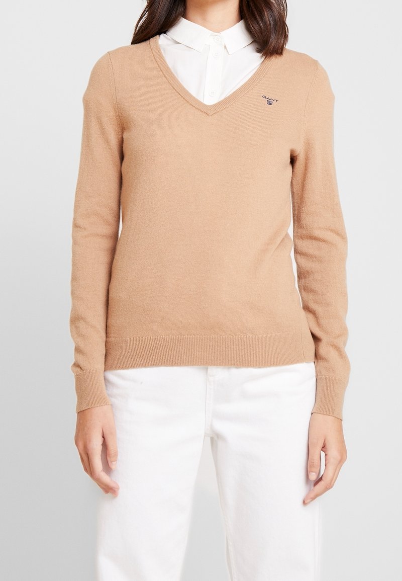 Personne portant un pull beige en V par-dessus une chemise blanche boutonnée avec un pantalon blanc, debout devant un fond gris clair uni.
