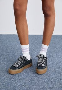 Czarne płócienne trampki z szaro-białym wzorzystym sznurowadłem, gumową podeszwą w kolorze brązowym, w połączeniu z białymi skarpetkami z purpurowymi paskami.