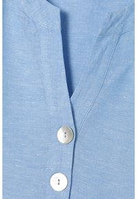 Camicia di lino azzurro chiaro con scollatura a V, caratterizzata da due bottoni rotondi di colore chiaro e una texture sottile. Cuciture visibili lungo i bordi.