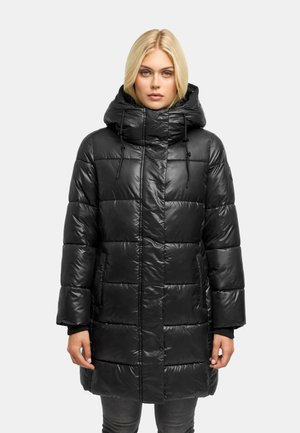 Femme blonde portant un manteau matelassé noir avec un col montant et une capuche, debout, les mains détendues le long du corps.