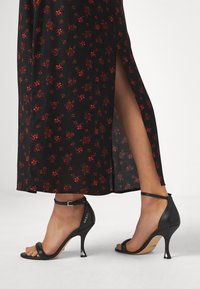 Schwarze High-Heelsandalen mit einem dünnen Knöchelriemen, offenen Zehen und einem markanten metallischen Absatz. Getragen mit einem floral gemusterten schwarzen Kleid.