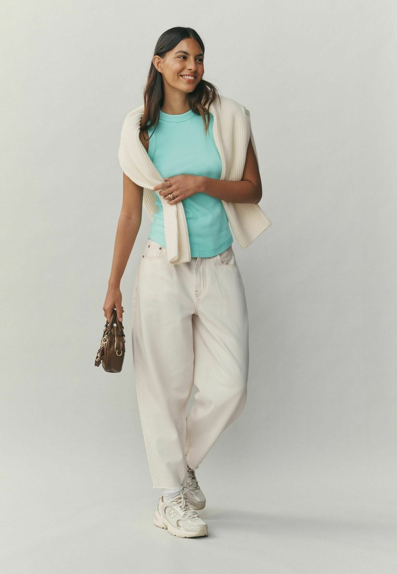 Débardeur ajusté bleu clair avec un cardigan crème côtelé posé sur les épaules, associé à un pantalon blanc ample et des baskets beiges. Petit sac à main marron.