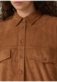 Eine Frau trägt ein schokoladenbraunes Wildlederhemd mit zwei Brusttaschen und sichtbar gewelltem Haar, das an den Kanten herausragt.