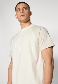 Lys beige t-skjorte med rund hals og korte raglanermer. Forsiden har en hvit "NEW YORK YANKEES" logo og subtil tekstur.