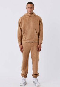 Felpa con cappuccio beige con tasca frontale e logo dorato, abbinata a pantaloni tapered coordinati e sneakers bianche con dettagli grigi.