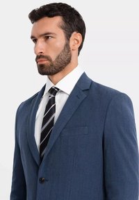 Niebieska marynarka z klapami w stylu notch, z jednopunktowym zapięciem i kieszenią na piersi. Noszona na białej koszuli z wzorzystym krawatem. Teksturowana tkanina.
