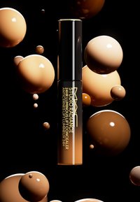 MAC Studio Radiance 24HR Luminous Lift Concealer i ett elegant svart rör, omgiven av glänsande bruna kulor mot en svart bakgrund.
