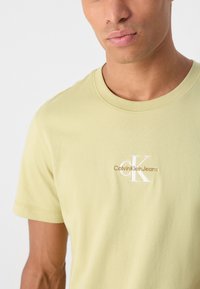 Gai zaļš kokvilnas T-krekls ar apaļu kakla izgriezumu, uz kura krūtīm ir izšūts "Calvin Klein Jeans" logo bēšā un baltā krāsā.