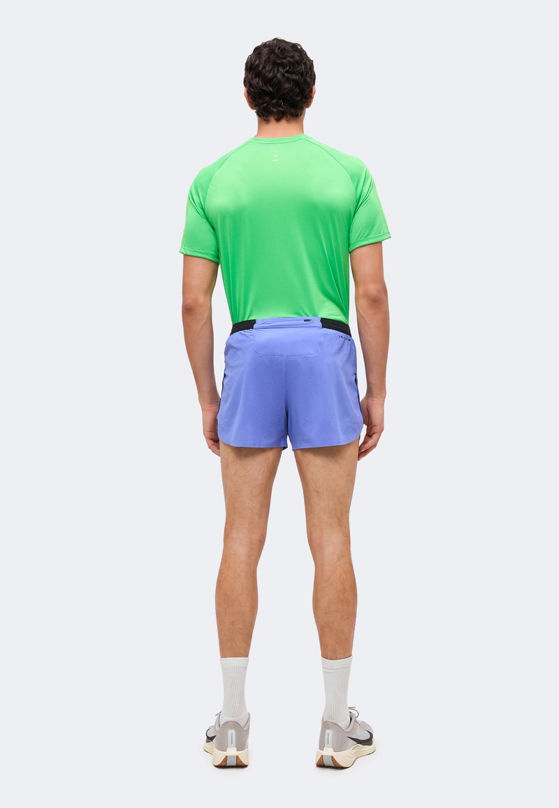Man standing facing away, wearing a green athletic shirt, light blue running shorts, white socks, and gray running shoes on a plain background.