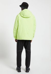 Hombre vistiendo una chaqueta con capucha verde lima, sombrero tipo cubo negro, pantalones negros con detalles reflectantes y zapatillas negras, de pie de espaldas.