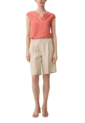 Femme portant un chemisier corail sans manches, un short beige taille haute longueur genou, et des sandales beiges à talons carrés avec brides, debout devant un fond blanc.