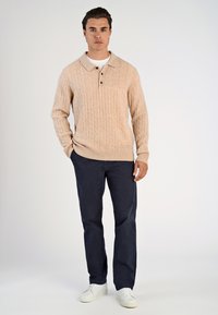 Beige kabelstickad tröja med krage och tre knappar, kombinerat med marinblå byxor och vita sneakers. Slät textur och avslappnad design.