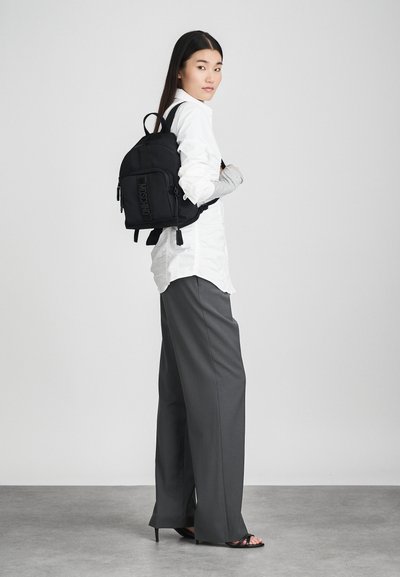 Γυναίκα ντυμένη με λευκό πουκάμισο και γκρι παντελόνι τύπου wide-leg, φορώντας μαύρο σακίδιο πλάτης και μαύρα ψηλοτάκουνα πέδιλα, μπροστά σε απλό γκρίζο φόντο.