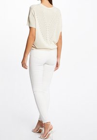 Kvinna som bär en krämfärgad stickad topp med kort ärm och perforerat V-mönster, vita åtsittande jeans och vita högklackade sandaler med remmar, står i bakifrån.