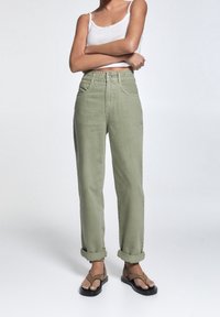 Mujer con pantalones de talle alto color verde oliva, remangados, una blusa sin mangas blanca y sandalias marrones, de pie con los brazos cruzados.