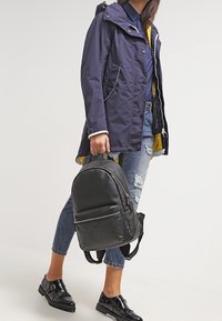 Black leather backpack with a rounded shape, front zip pocket, and adjustable straps. Model wears a navy waterproof jacket with yellow lining.