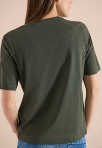 Femme portant un t-shirt à manches courtes vert olive, vue de dos, associée à un jean en denim bleu sur un fond neutre.