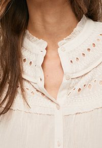 Camicia bianca con bottoni sul davanti, caratterizzata da un colletto arricciato, dettagli ricamati e motivi intagliati lungo il décolleté.